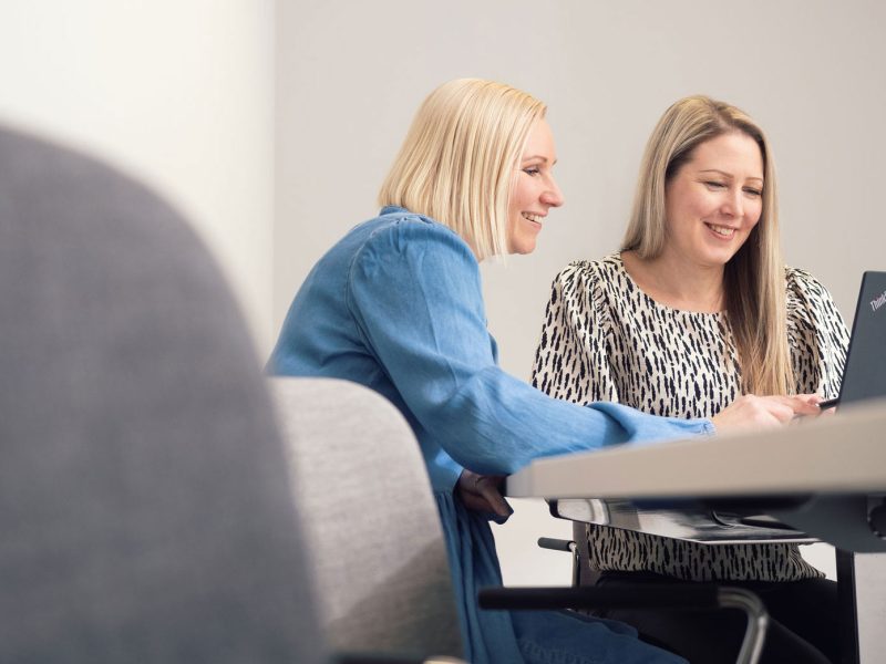 Kaksi naisoletettua henkilöä istumassa hymyillen kannettavan tietokoneen ääressä valkoisessa toimistotilassa. Toisella henkilöllä on vaaleat polkkamittaiset hiukset ja sininen tunika. Toisella henkilöllä on vaaleanruskeat pidemmät hiukset, mustavalkoinen pusero.