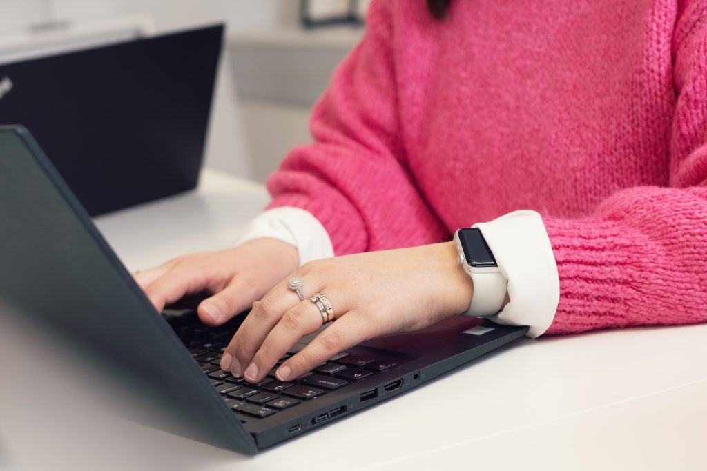 Lähikuvassa pinkkiin paitaan pukeutuneen henkilön kädet näppäilemässä mustan kannettavan tietokoneen näppäimistöä. Vasemman käden ranteessa on älykello ja nimettömässä ja keskisormessa sormukset.