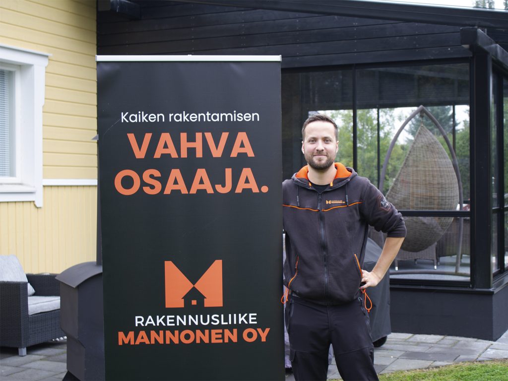 Rakennusliike Mannonen Oy:n toimitusjohtaja Joona Mannonen seisoo keltaisen talon ja mustan terassin edustalla. Vieressä roll-up, jossa teksti 
